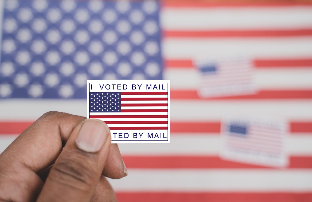 Early ballot "I voted by mail" sticker held up against an American flag background.