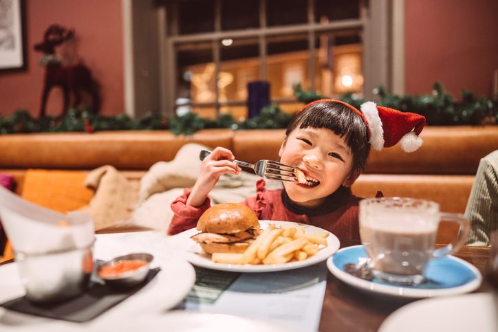 Lovely little girl in Santa hat headband smiling joyfully at the camera while enjoying burger & chips in a restaurant.