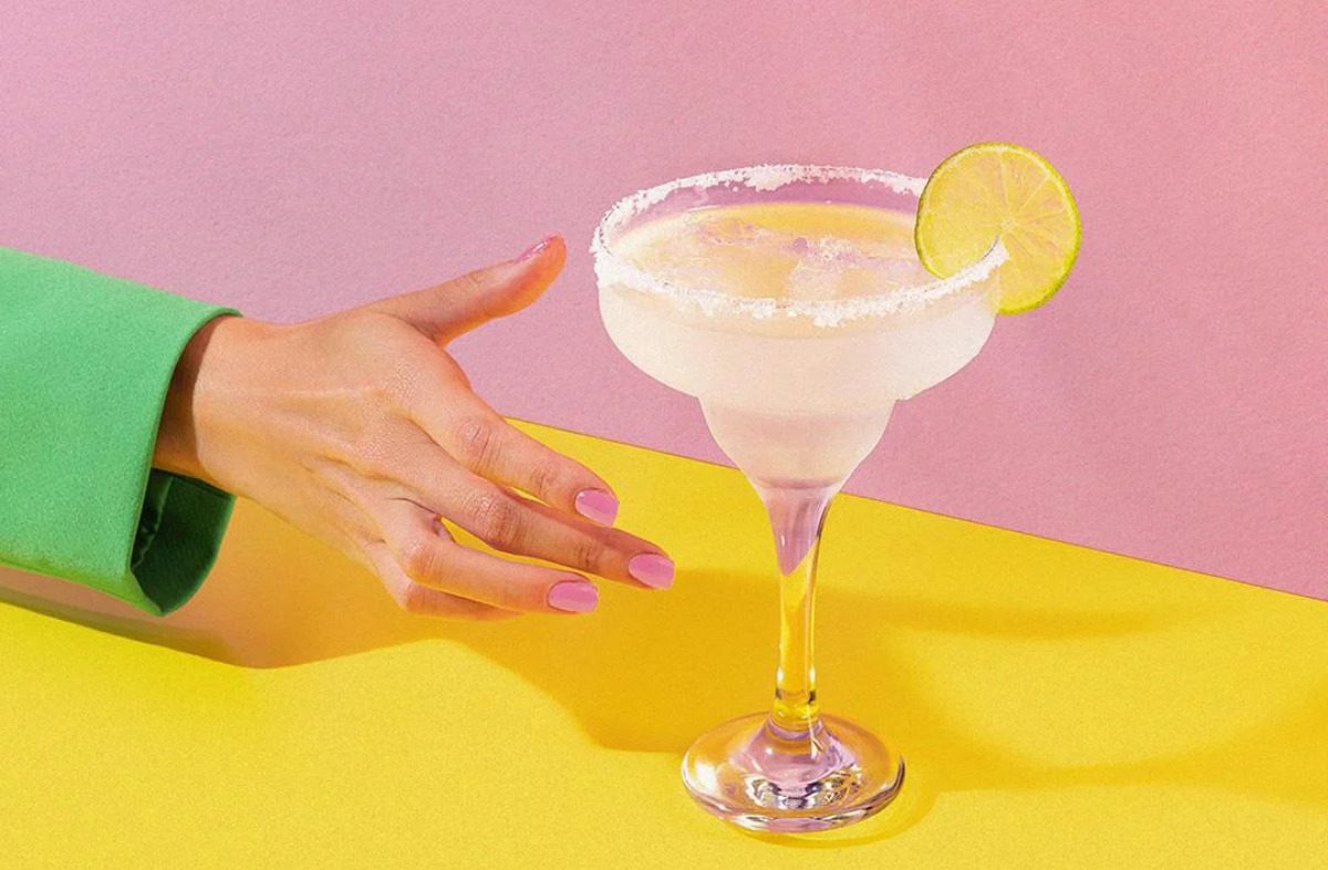 Woman's hand reaching for an alcoholic drink against a pink and yellow background.